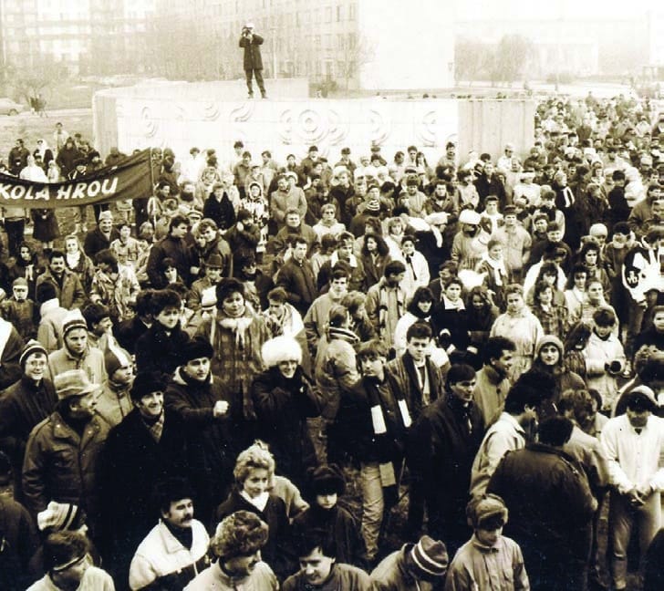 Dav na zhromaždení pri dome kultúry, na betónovom poloblúku v pozadí stojí muž s kamerou (zvýraznený krúžkom)